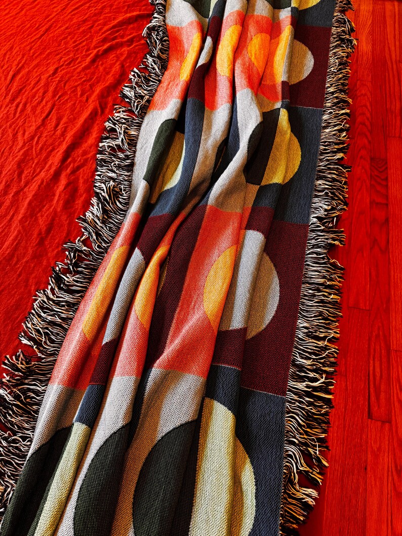 May include: A colorful throw blanket with a geometric pattern of circles and rectangles in shades of orange, yellow, gray, green, and navy blue. The blanket has a fringed edge and is draped over a red surface.