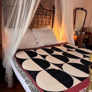 May include: A black and white geometric patterned throw blanket with a burgundy border and fringe. The blanket is on a bed with a white canopy and white pillows.