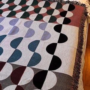 May include: A woven throw blanket with a geometric pattern of half circles in shades of red, green, blue, and black. The blanket has a fringe border.