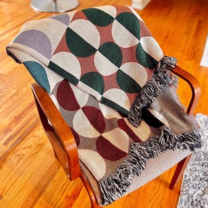 May include: A woven throw blanket with a geometric pattern of green, white, and brown half circles. The blanket is draped over a wooden chair with a light brown fabric seat.
