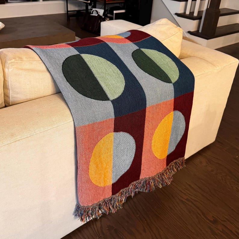 May include: A colorful woven throw blanket with a geometric pattern of circles and squares. The blanket features a mix of colors including blue, green, red, orange, and gray. The throw has a fringed edge and is draped over a cream-colored sofa.
