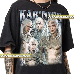 May include: Black t-shirt featuring a graphic print of four figures with white hair against a dark background. The text "KAR'NISS" is printed in blue and white lettering at the top of the graphic. The text "MIDAGEN" is printed in white lettering at the top right corner of the graphic.