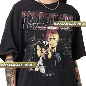Puede incluir: Camiseta negra con el texto "RESIDENT EVIL DEAD AIM" en rojo y blanco. El gráfico incluye imágenes de personajes con armas. La camiseta tiene un aspecto vintage y desgastado.