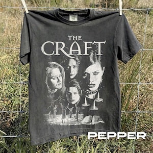 May include: A charcoal grey short-sleeve t-shirt featuring a white graphic print of four faces and the text THE CRAFT. The design includes silhouettes of figures with raised arms, lit candles, and an open book at the bottom of the shirt.