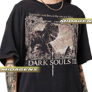 May include: Black t-shirt featuring a distressed graphic from the video game Dark Souls III. The design includes a knight, a sword, and the game's title. The shirt also has the text "Surrender your fires, to the one true Heir."