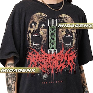 Może przedstawiać: Czarny t-shirt z grafiką przedstawiającą dwie twarze zombie i zieloną fiolkę. Projekt zawiera czerwony tekst i słowa "YOU ARE DEAD". Koszulka ma postarzany wygląd. Koszulka jest prawdopodobnie wykonana z bawełny.