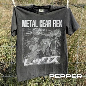 May include: A charcoal gray short-sleeve t-shirt featuring a white graphic print of the Metal Gear Rex robot. The design includes the text METAL GEAR REX in English and Japanese characters, with a vertical column of Japanese text on the left side.