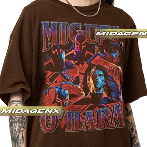 Puede incluir: Camiseta marr&oacute;n con un estampado gr&aacute;fico vintage de un grupo de personas, incluyendo un hombre con el nombre "MIGUEL O'HARA" impreso en letras grandes. El gr&aacute;fico est&aacute; en tonos de rojo, azul y negro.