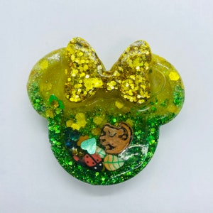 May include: A yellow and green Minnie Mouse-shaped resin accessory with a glittery bow. The design includes a cartoon lion, ladybug, and leaf embellishments. The accessory is filled with green glitter.