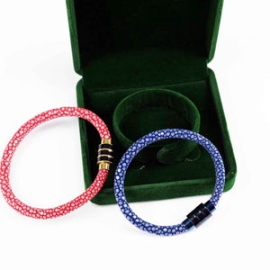 Könnte beinhalten: Zwei Lederarmbänder mit Magnetverschlüssen. Ein Armband ist rot mit einem goldenen Akzent und das andere ist blau mit einem silbernen Akzent. Die Armbänder befinden sich in einer grünen Samtbox.