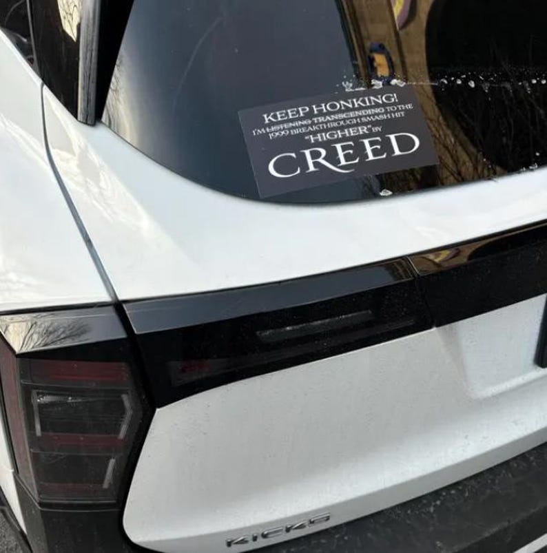 May include: A white car with a black bumper and a sticker on the back window that reads "KEEP HONKING! I'M LISTENING TRANSCENDING TO THE 1999 BREAKTHROUGH SMASH HIT "HIGHER" BY CREED"
