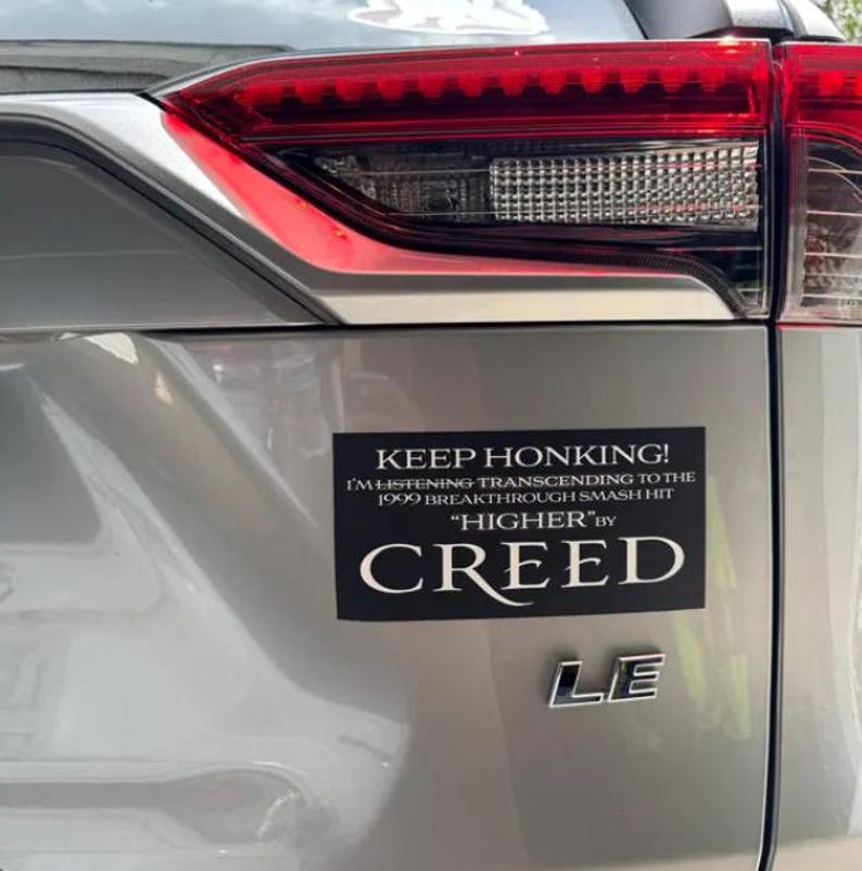 May include: A black and white bumper sticker on a silver car that reads "KEEP HONKING! I'M LISTENING TRANSCENDING TO THE 1999 BREAKTHROUGH SMASH HIT "HIGHER" BY CREED"