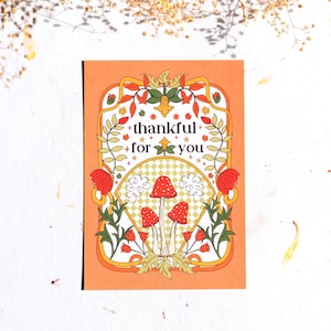 May include: A peach-colored card with a decorative border featuring flowers and mushrooms. The text "thankful for you" is written in black lettering.