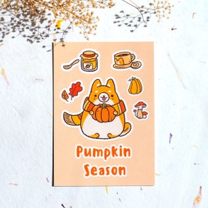 May include: A cute orange and white dog wearing a scarf holds a pumpkin and is surrounded by fall-themed stickers, including a cup of coffee, a jar of honey, a mushroom, and an acorn. The text "Pumpkin Season" is written below the dog.