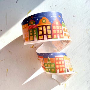 May include: A roll of washi tape with a festive Christmas town design. The tape features colorful houses with windows, a starry night sky, and a green tree.
