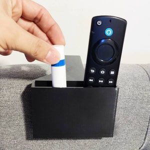 Puede incluir: Un soporte de control remoto negro con un compartimento para un mando a distancia y un tubo pequeño. El soporte está unido a un sofá gris.