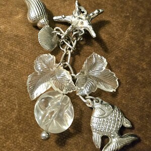 May include: A silver-toned charm bracelet pendant featuring marine life and botanical elements. The pendant includes a starfish, fish, seashells, and clear leaf and orb-shaped charms, all connected by a chain.