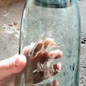 May include: A vintage Ball Mason jar with a silver lid. The clear glass jar has embossed lettering that reads "Ball Mason". The jar is held by a person. The jar is a light blue-green color.