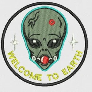 May include: A round, green alien head with black eyes and a red kiss mark on its forehead. The alien is wearing a silver collar with a red ball attached. The alien is surrounded by a turquoise circle with the words "Welcome to Earth" written in black.