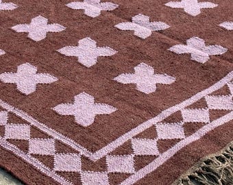 Puede incluir: Una alfombra rectangular con una base de color marrón oscuro y un patrón repetitivo de formas de cruz en color lavanda claro. La alfombra incluye un borde con un motivo de diamantes y acabados con flecos largos, finos y marrones en los extremos.