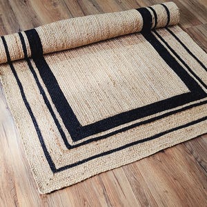 May include: A natural jute rug with a rolled-up section. The rug features a beige background with a black border and stripes. The rug is rectangular and appears to be made of woven fibers, suitable for home decor.