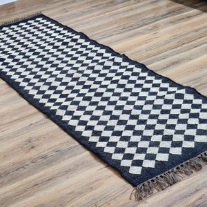 Op de afbeelding: Zwart-wit geblokt vloerkleed met franjes aan de randen. Het vloerkleed is gemaakt van geweven materiaal en heeft een geometrisch patroon.