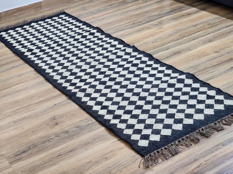 Op de afbeelding: Zwart-wit geblokt vloerkleed met franjes aan de randen. Het vloerkleed is gemaakt van geweven materiaal en heeft een geometrisch patroon.