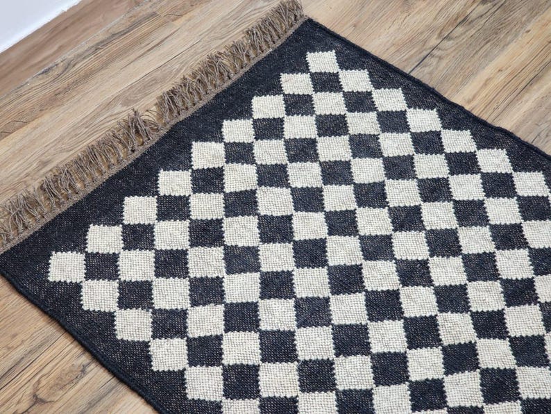 Op de afbeelding: Zwart-wit geblokt vloerkleed met franjes aan de randen. Het vloerkleed is gemaakt van geweven materiaal en heeft een geometrisch patroon.