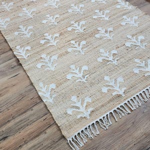 May include: A natural fibre rug with a white floral pattern and fringe edging. The rug is laid out on a wooden floor.