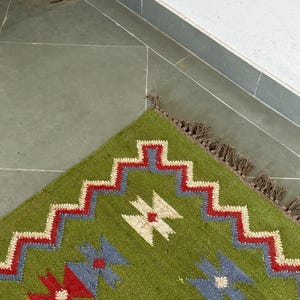 May include: A corner of a green rug with a geometric pattern. The rug features a zigzag border in red and white, with blue and red floral-like shapes. The rug has a fringe edge.