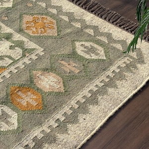 Può includere: Un tappeto intrecciato con uno sfondo verde salvia e un motivo ripetuto di forme a diamante nei colori giallo senape, crema e marrone chiaro. Il tappeto presenta un bordo a zigzag e frange marrone scuro lungo il lato corto.