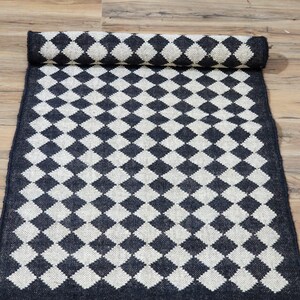 Op de afbeelding: Zwart-witte ruitvormige vloerkleed met franjes. Het vloerkleed is gemaakt van natuurlijke vezels en heeft een rustieke uitstraling.