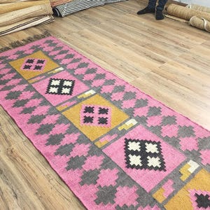 May include: A rectangular pink and gray patterned rug with yellow and black square designs. The rug has a fringe on the short ends and is placed on a wooden floor. Rolled rugs are visible in the background.