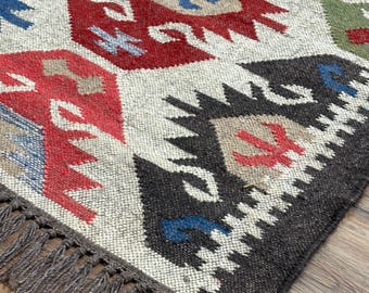 May include: Close-up of a woven rug with a geometric pattern. The rug features a cream-colored background with red, blue, black, and green geometric shapes. The rug has a fringe along one edge.