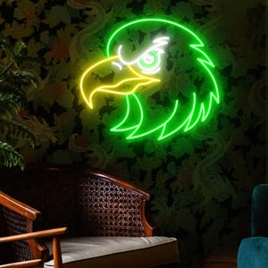 May include: A neon sign in the shape of an eagle's head, with a green outline and yellow beak. The sign is mounted on a dark floral wallpaper background. The eagle's eye is white.