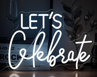 May include: A white neon sign that reads "LET'S Celebrate" in a cursive font. The sign is on a clear acrylic backing and sits on a wooden surface. A wine glass and decorative plant are in the background.