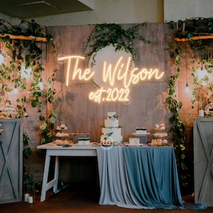 May include: A wedding reception dessert table featuring a neon sign that reads "The Wilson est. 2022". The table is decorated with greenery and has a white cake, cupcakes, and other desserts.