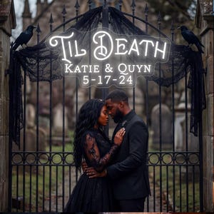 May include: A couple stands before a wrought iron gate, a neon sign above reading "Til Death." The sign also displays "Katie & Quyn" and the date "5-17-24." Black crows sit on the gate, with black fabric draped from the top. The couple is dressed in black.