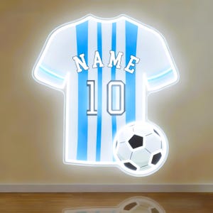 May include: A white and light blue soccer jersey-shaped wall decoration with the name "NAME" and the number "10". A soccer ball is positioned below the jersey. The design has a glowing outline.