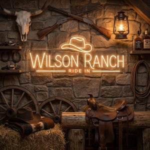 May include: A rectangular neon sign displaying the text WILSON RANCH RIDE IN with a cowboy hat icon above the lettering. The sign is mounted on a stone wall decorated with a steer skull, crossed rifles, a lantern, a saddle, and a cowboy hat.