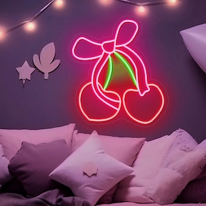 May include: A neon sign shaped like cherries with a bow, outlined in bright pink with a green stem. It's mounted on a dark purple wall above a bed with pillows. The scene is illuminated by fairy lights.