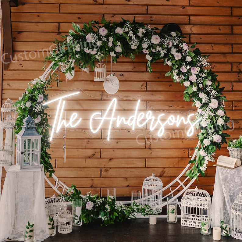May include: White neon sign with the text "The Andersons" in a cursive font, surrounded by a white metal hoop decorated with greenery and white flowers.