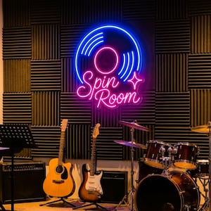 Könnte beinhalten: Ein Neonschild mit dem Schriftzug Spin Room in pinker Schreibschrift und einer blauen Schallplattengrafik darüber. Das Schild ist an einer dunkelgrauen Akustikschaum-Wand montiert, über einem Musikstudio-Setup mit Gitarren und Schlagzeug.