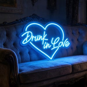 Op de afbeelding: Een blauw neonbord in de vorm van een hart met de tekst "Drunk in Love" in een sierlijk lettertype. Het bord staat op een grijze fluwelen bank met sierlijke details.