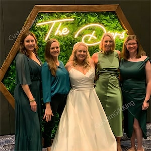 May include: Five women stand in front of a green moss wall with a wooden hexagonal frame. The wall has a neon sign that reads "The D's". The women are wearing green dresses and the woman in the centre is wearing a white wedding dress.