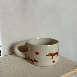 Könnte beinhalten: Cremefarbene Keramiktasse mit braunen Dackel-Illustrationen und roten Blumenakzenten. Die Tasse hat einen abgerundeten Henkel und steht auf einem hellen Holzregal. Ein bezauberndes und skurriles Design.