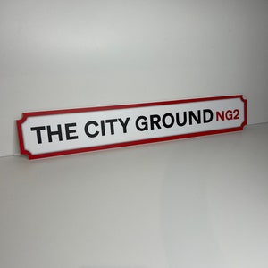 Op de afbeelding: Wit bord met rode rand en zwarte tekst die "THE CITY GROUND NG2" luidt.