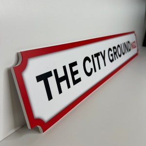 Op de afbeelding: Een wit bord met een rode rand en de tekst "THE CITY GROUND" in zwarte letters.