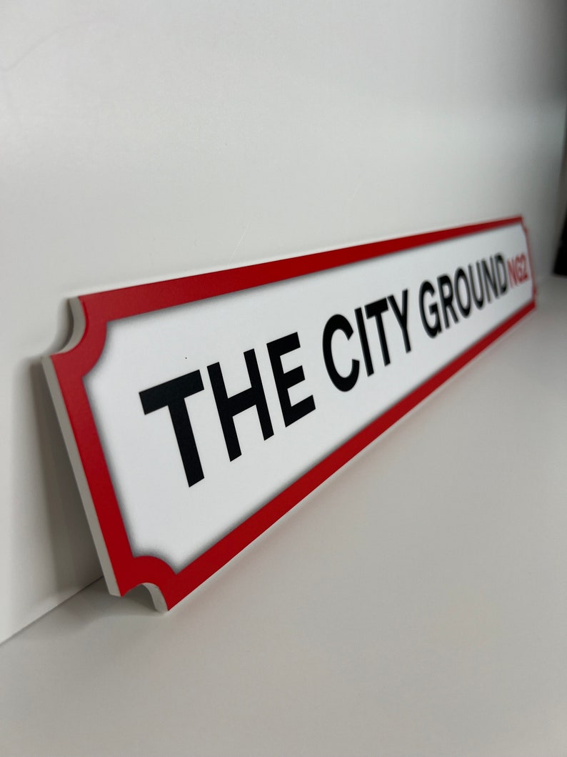 Op de afbeelding: Een wit bord met een rode rand en de tekst "THE CITY GROUND" in zwarte letters.