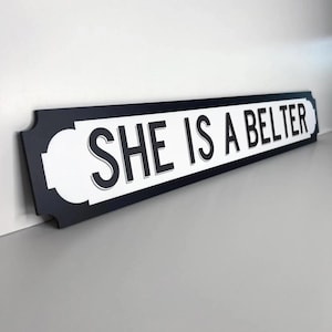 May include: A black and white sign with the text "SHE IS A BELTER". The sign has a black border and white interior with rounded corners. The text is in a bold, sans-serif font. A decorative item.
